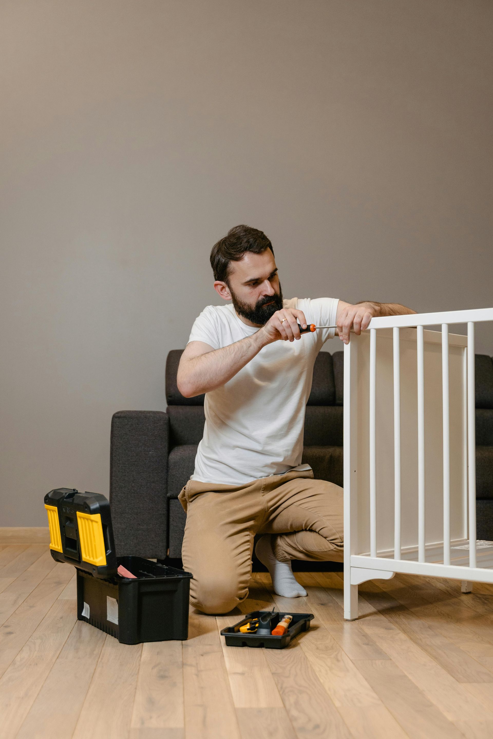 Man assembling a white crib on a wooden floor, with a toolbox nearby.