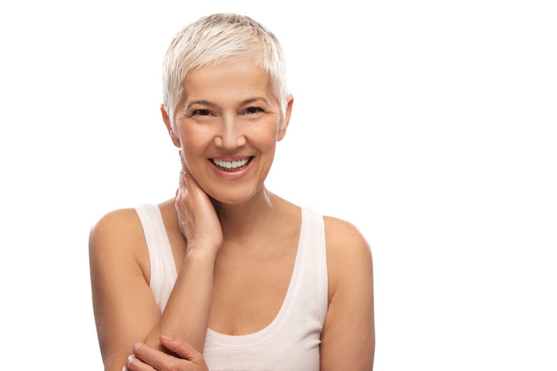 A woman with short gray hair is smiling and touching her face.