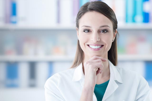 A female pharmacist is smiling with her hand on her chin in a pharmacy.