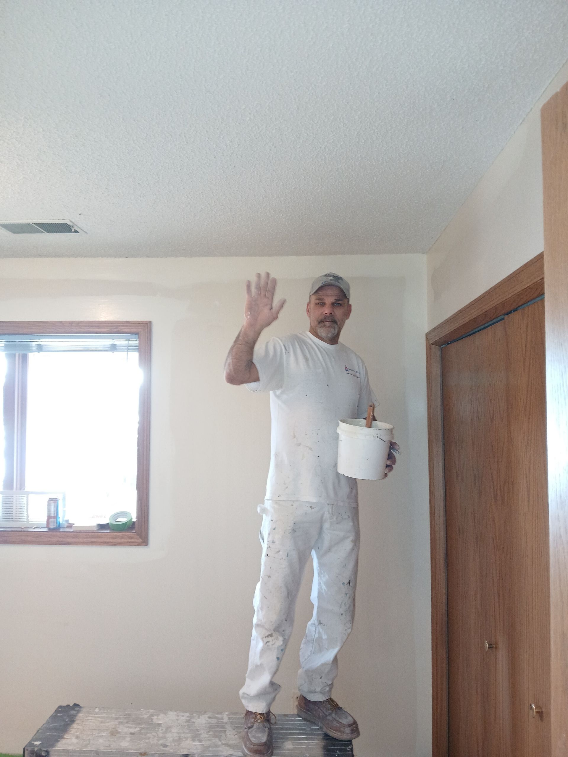 A man is standing on a ladder in a room holding a bucket of paint.