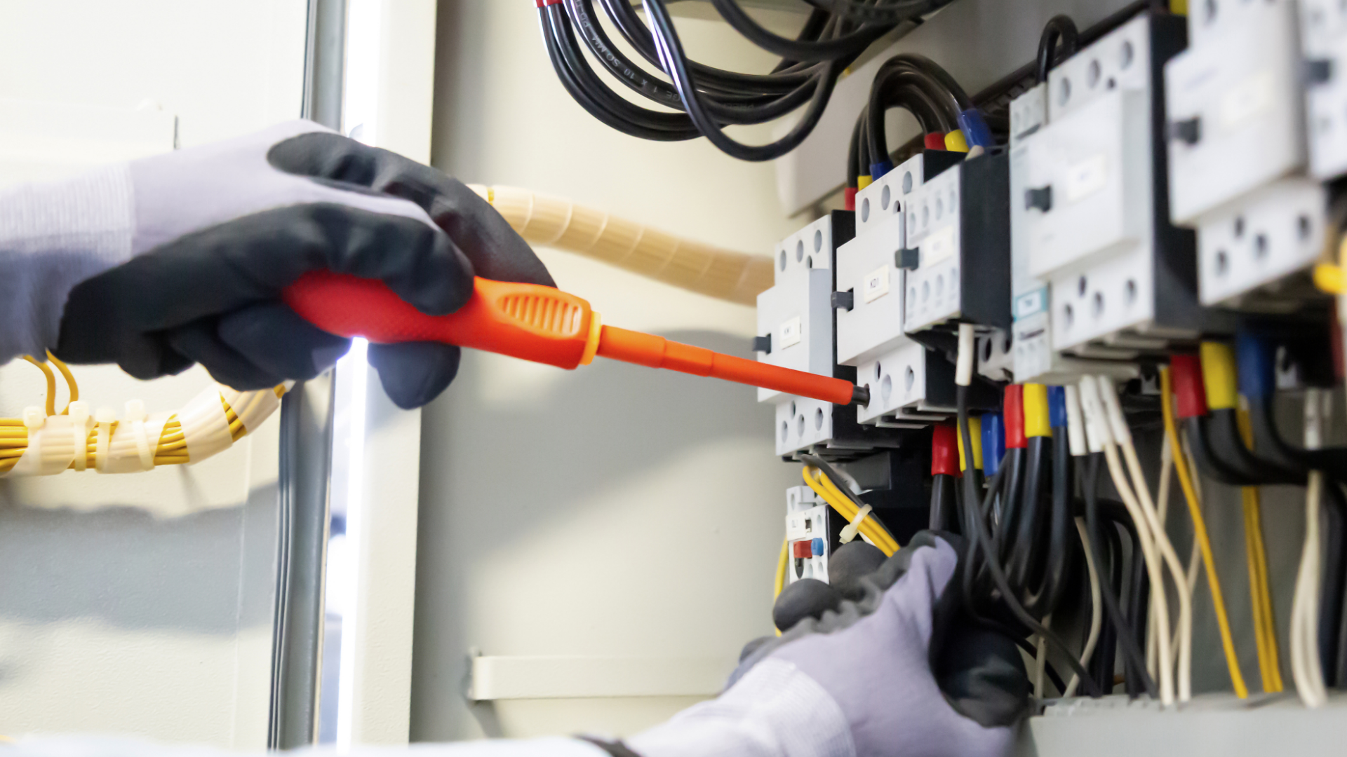 Person wearing gloves using a screwdriver on electrical panel, connecting wires.