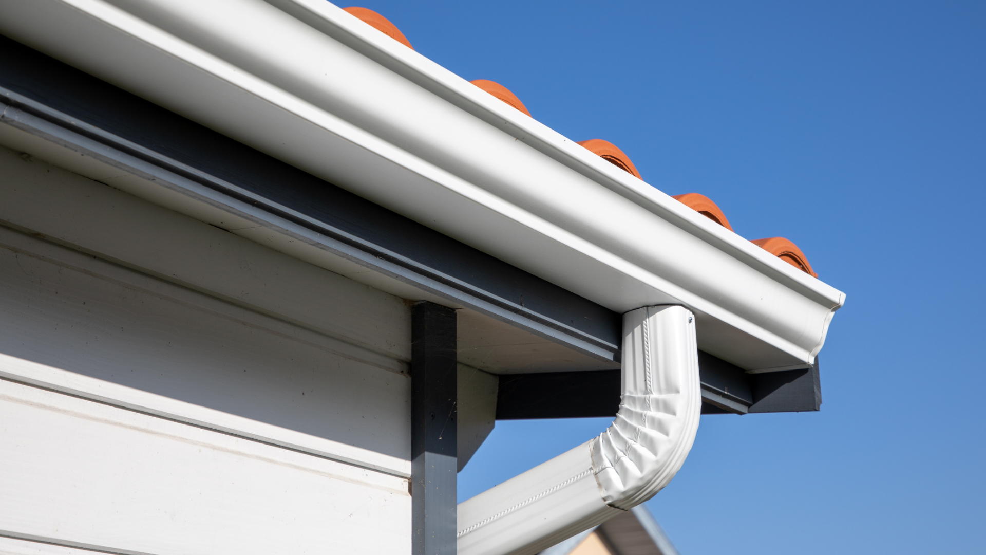 White rain gutter and downspout on a building with an orange tile roof against a clear blue sky.