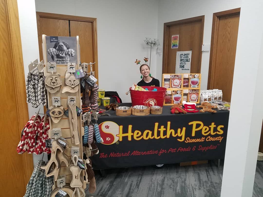 A vendor booth for Healthy Pets, featuring a display rack of pet accessories and a table with treats in a room with doors.