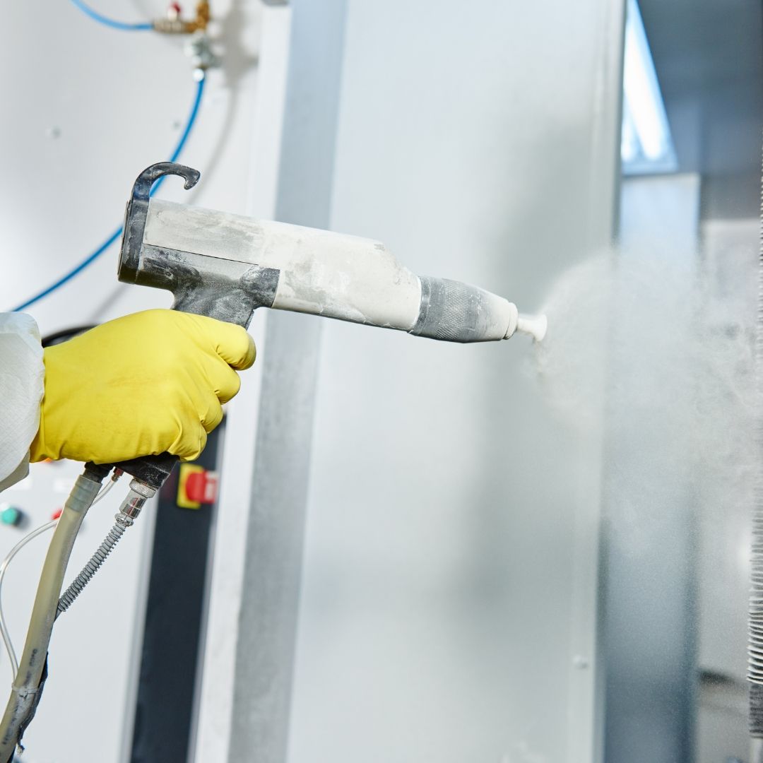 Person spraying powder coating with a handheld gun. Yellow glove, gray gun, and white powder.