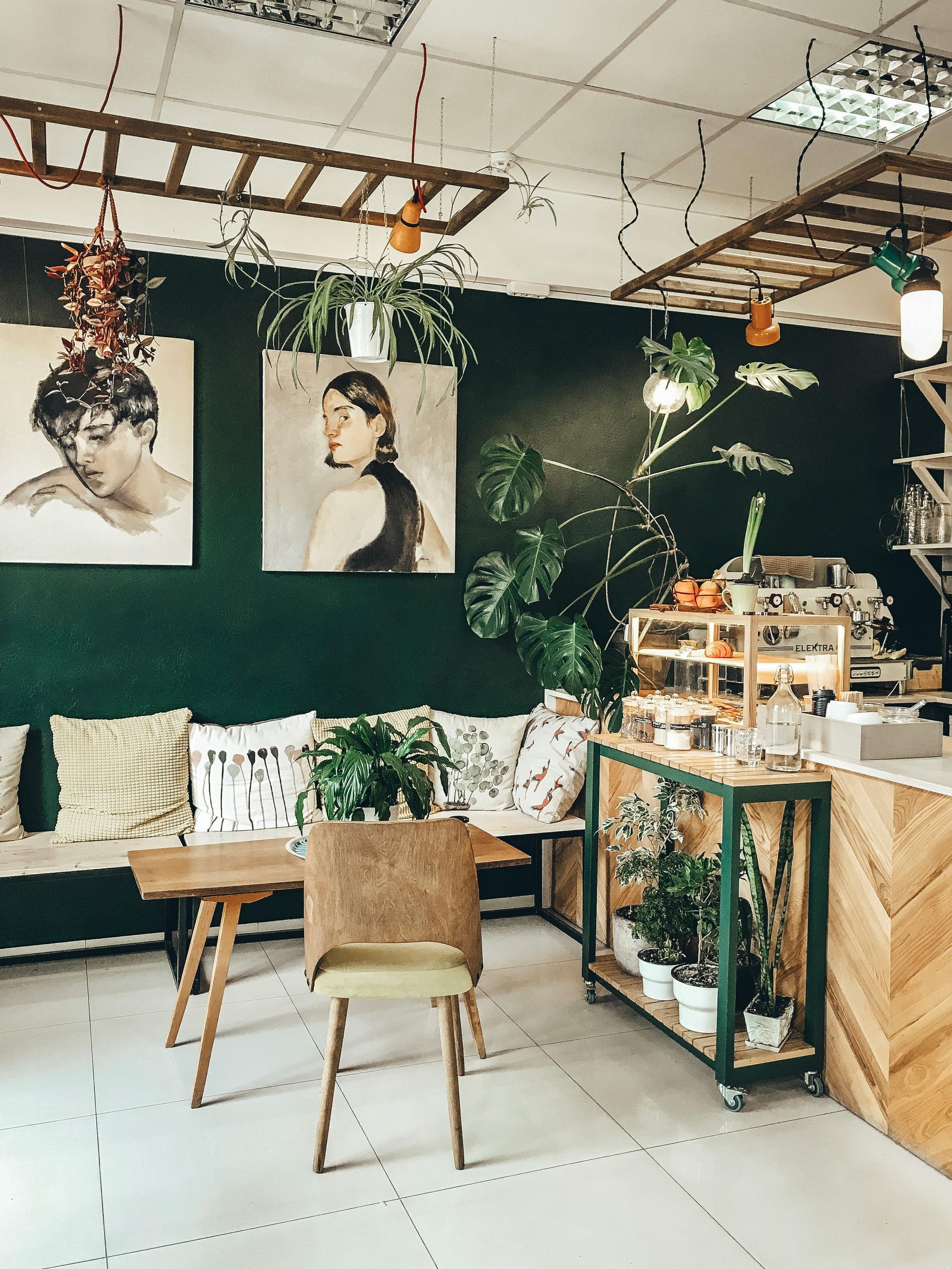 Cozy cafe interior with dark green walls, art, and plants. Wooden furniture, counter, and hanging lights.