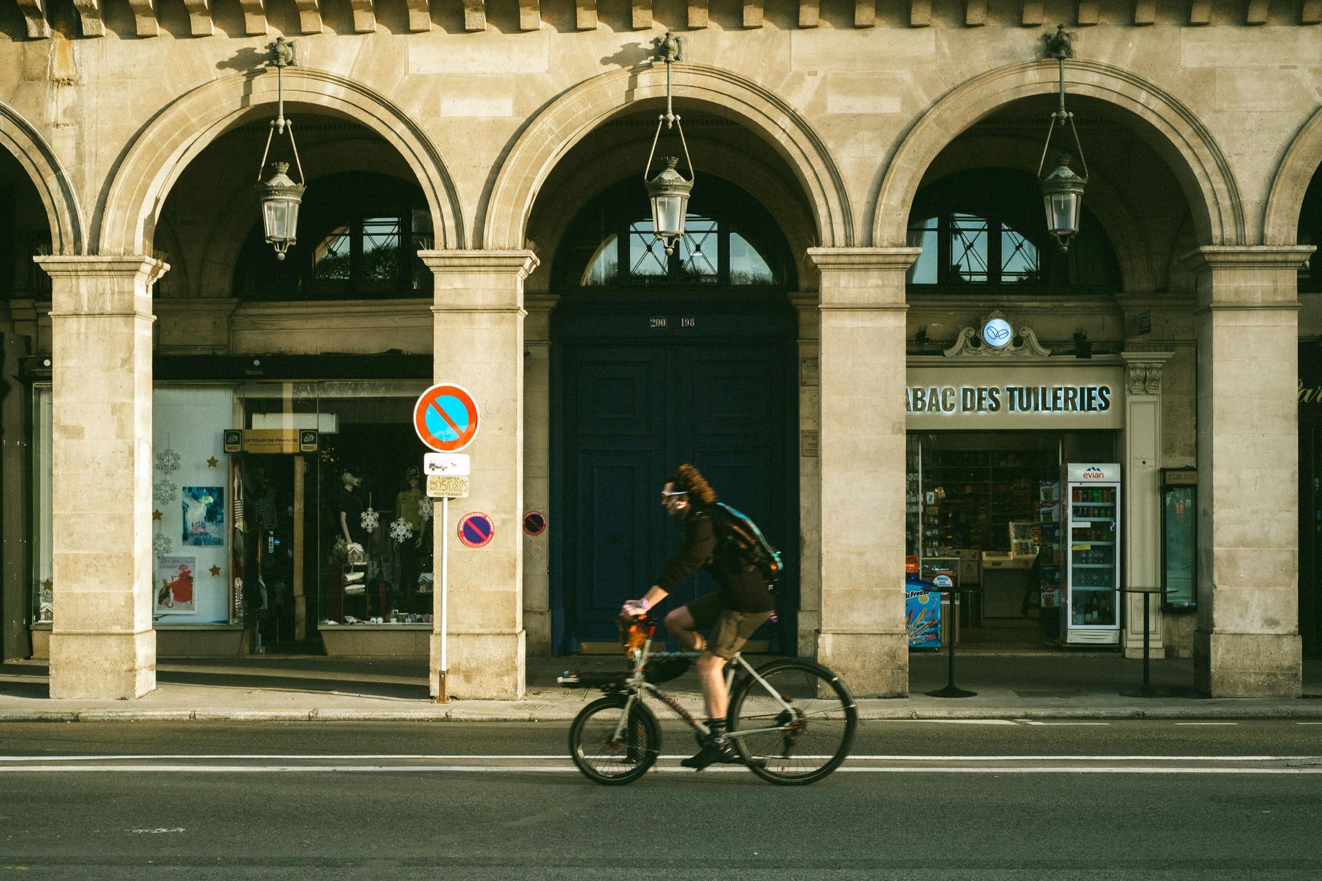 Person riding bicycle on street under arched stone arcade. Shops in background.