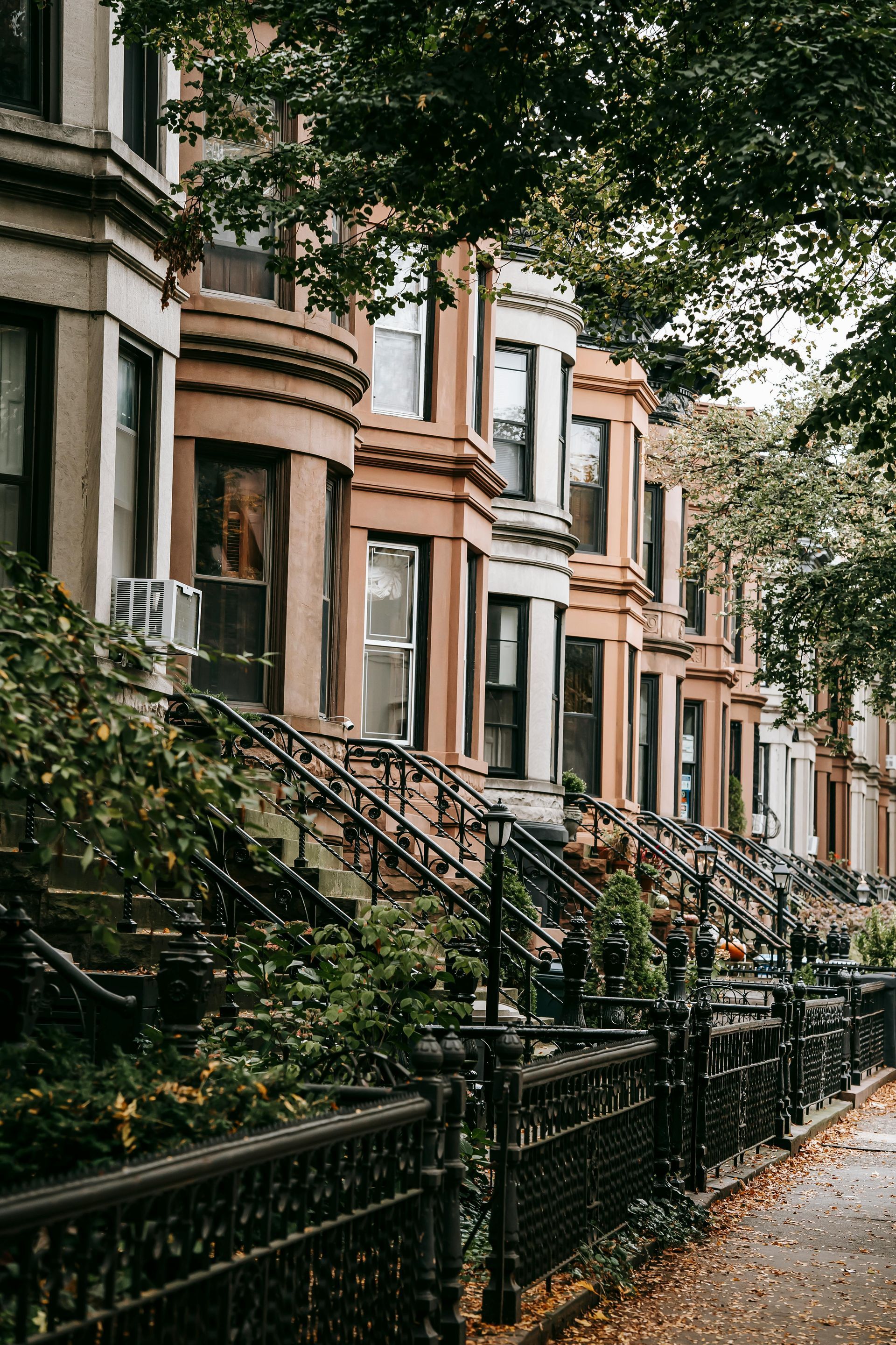 Row of brownstone buildings with bay windows, wrought iron fences, and leafy trees.