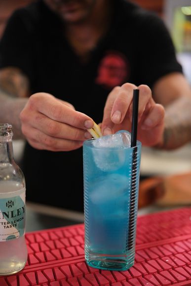 Il barista guarnisce un cocktail blu in un bicchiere alto con ghiaccio e una cannuccia, accanto a una bottiglia di soda.