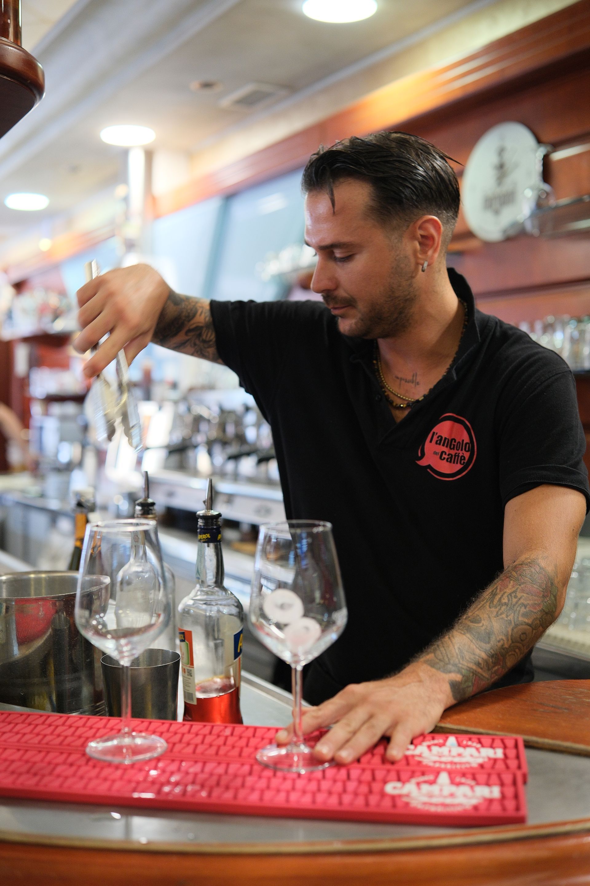 Barista che mescola un drink dietro al bancone; bicchieri, ghiaccio, tappetino rosso; braccia tatuate; interno.