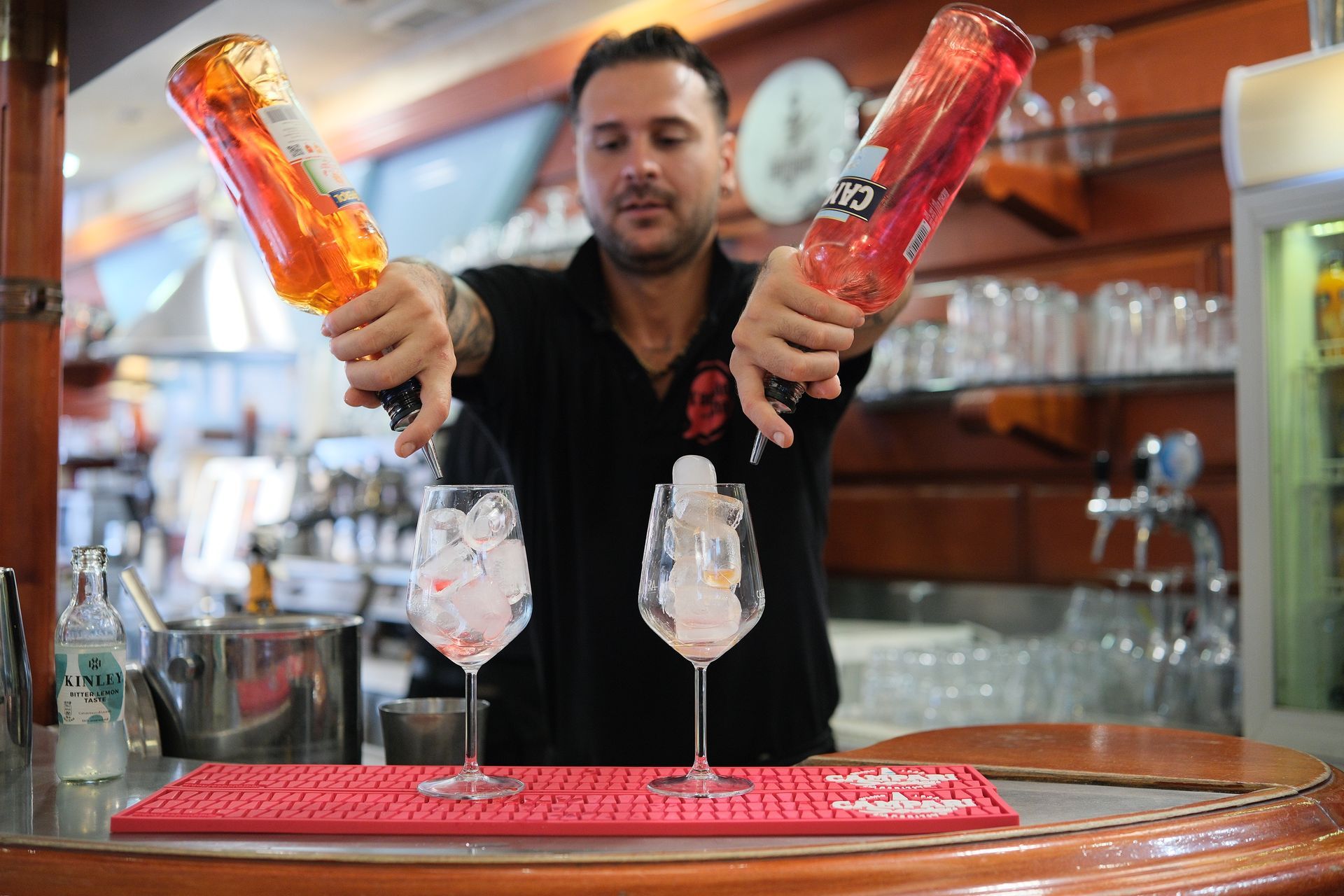 Barista che versa liquori colorati nei bicchieri con ghiaccio; dietro un bancone.