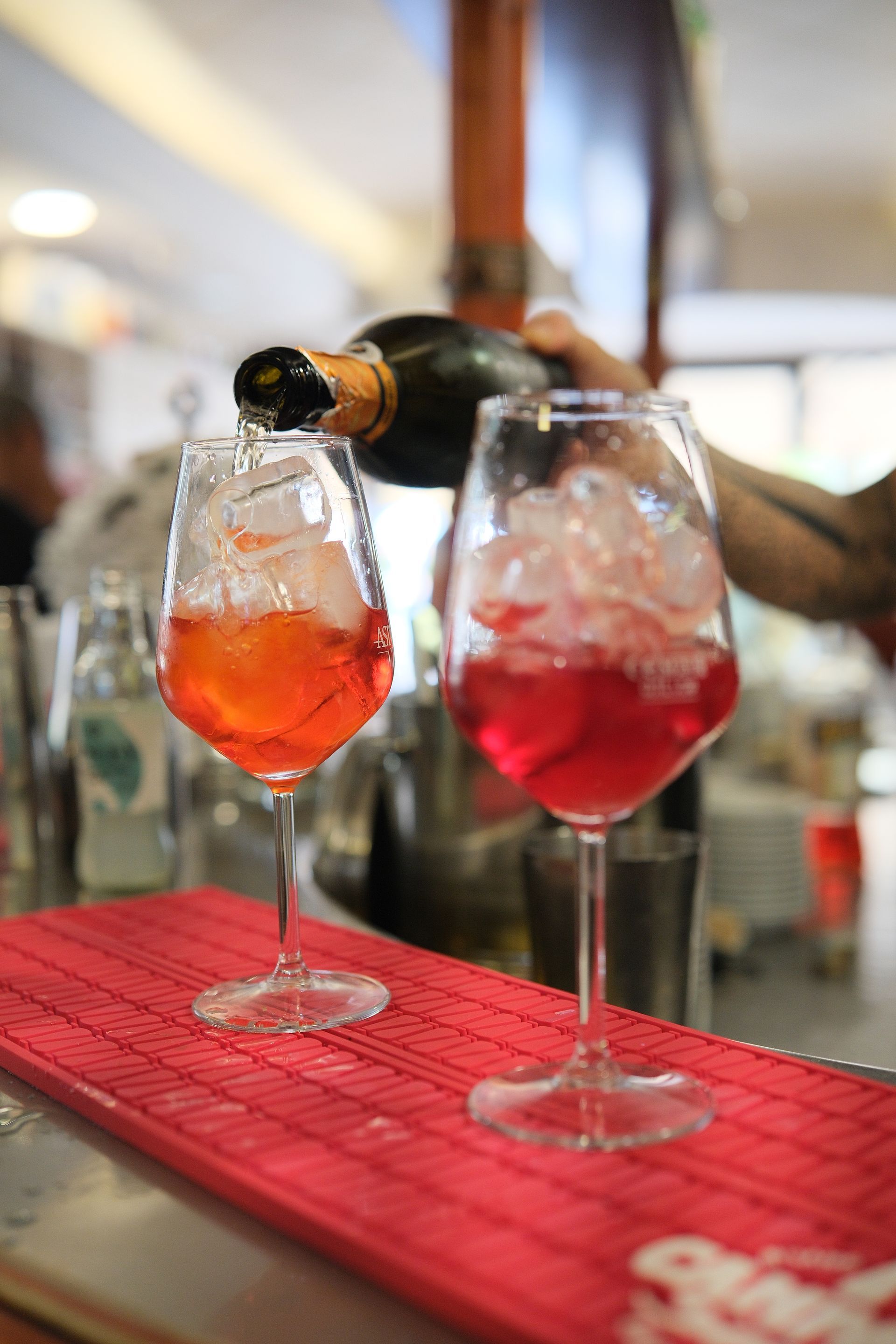 Un barista versa champagne in un bicchiere di Aperol Spritz con ghiaccio, su un sottobicchiere rosso.