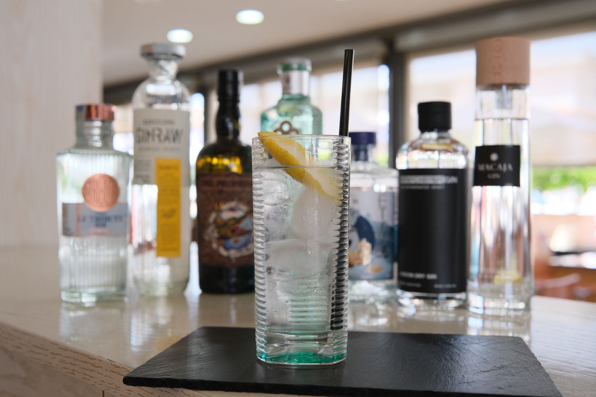 Cocktail gin tonic con una fetta di limone e una cannuccia nera, circondato da varie bottiglie di gin su un bancone.