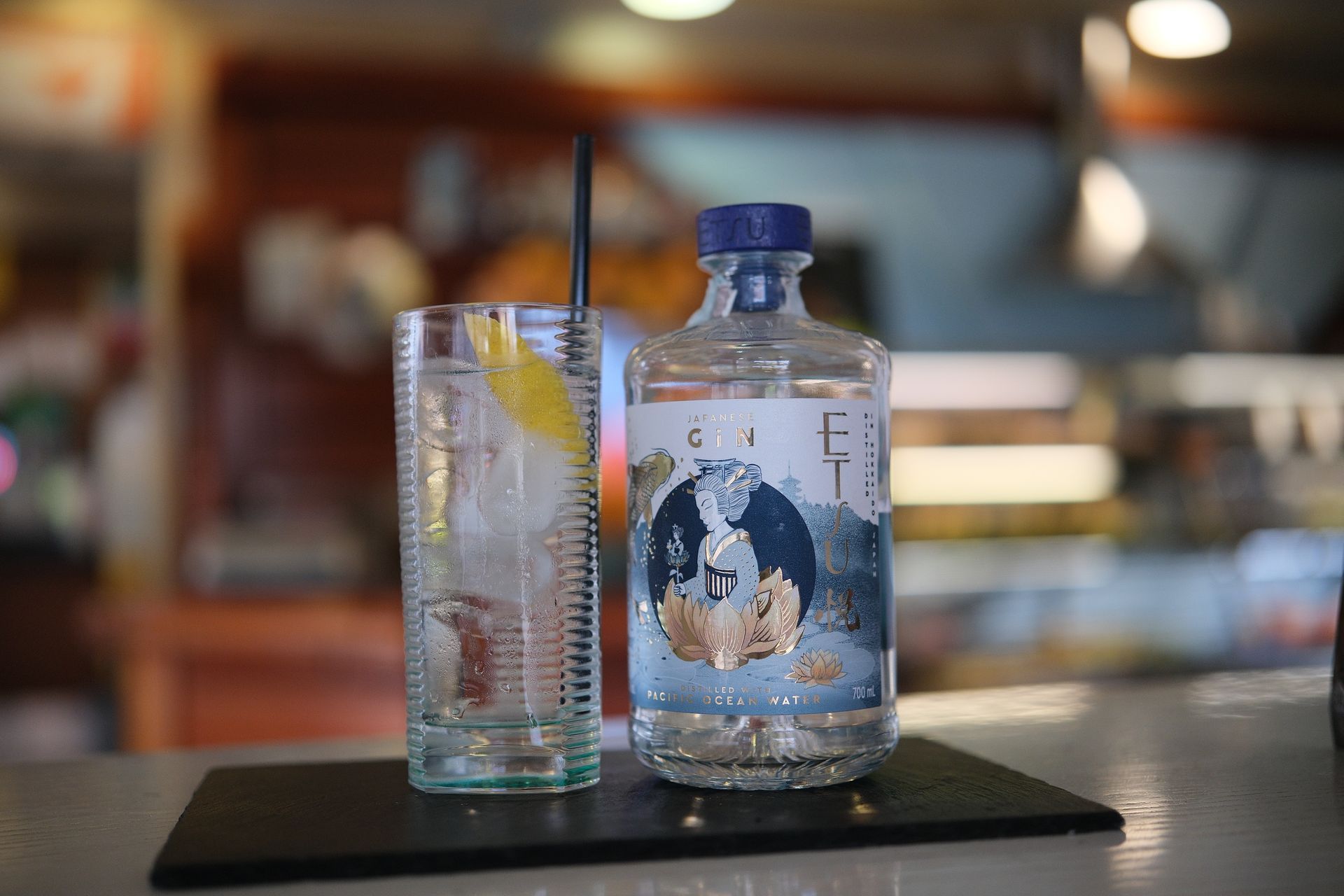 Bottiglia di gin e cocktail su un bancone del bar; la bottiglia è blu e oro, il drink ha limone e cannuccia.