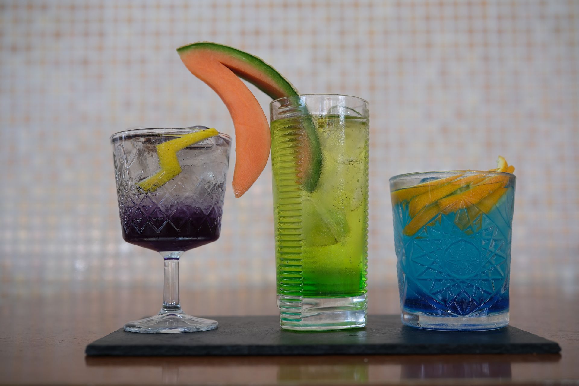 Tre cocktail colorati su un vassoio nero, guarniti con frutta e scorza, su uno sfondo piastrellato.