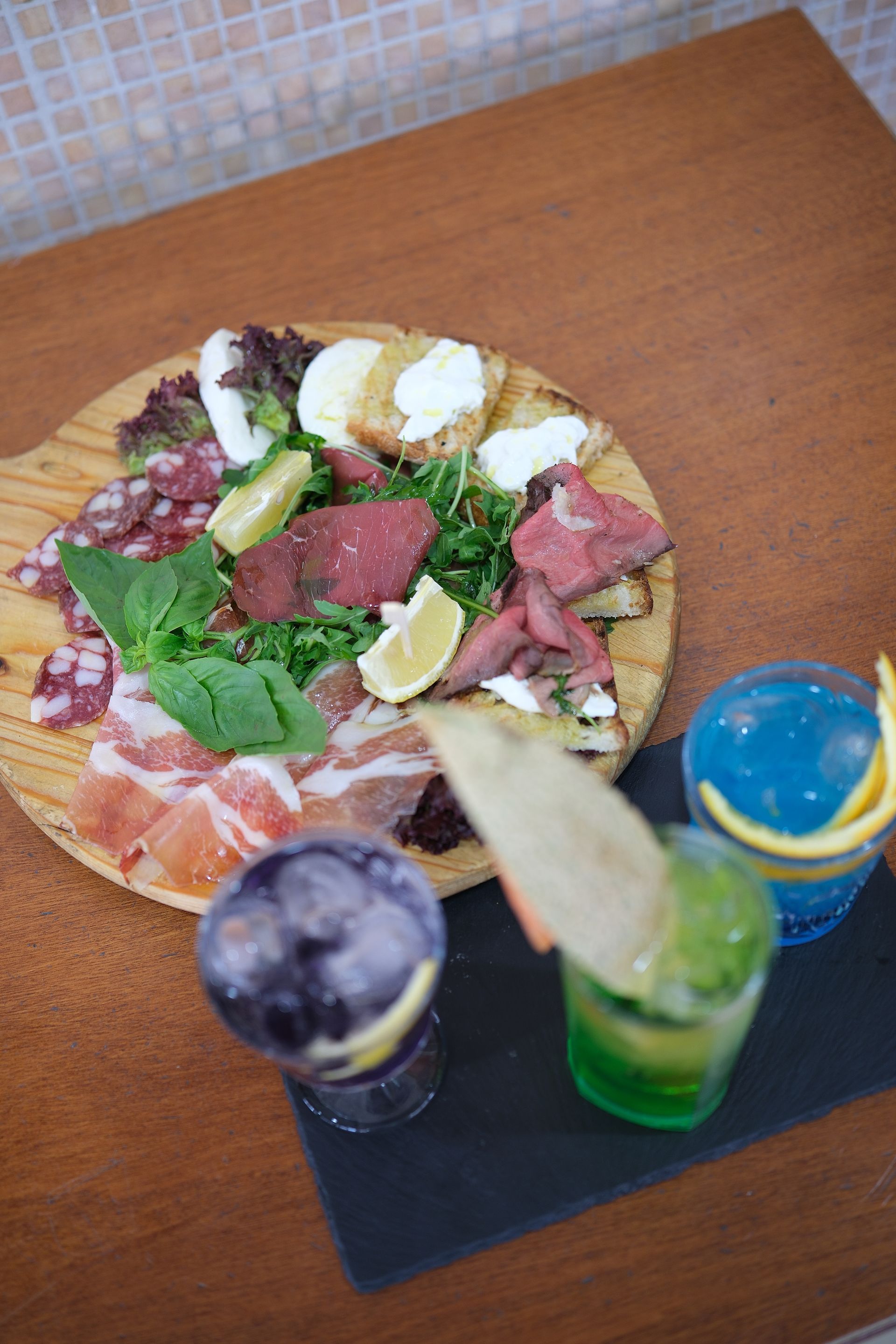 Tagliere di legno con salumi, formaggi, verdure e tre bevande colorate su un tavolo.