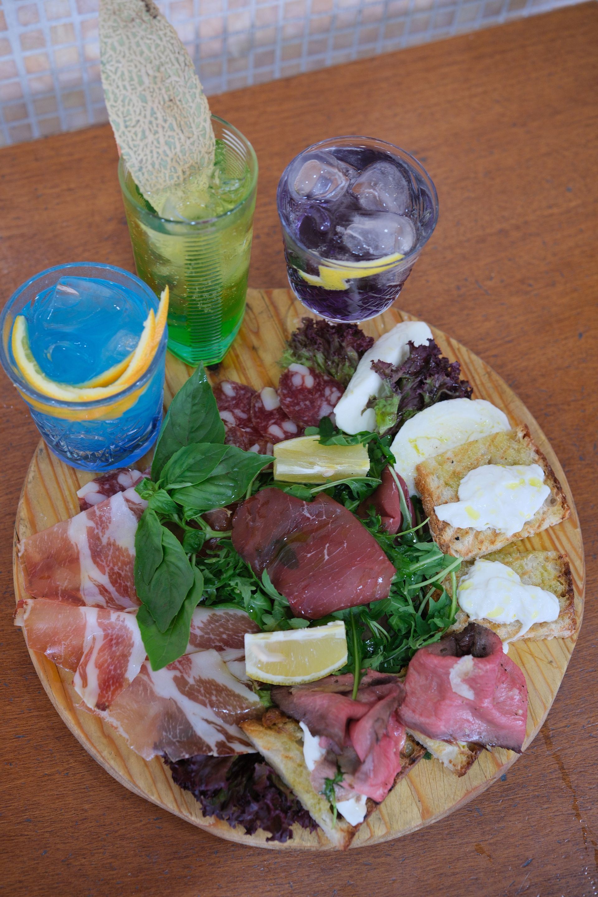 Tagliere di salumi con carne, formaggio, verdure, spicchi di limone e tre cocktail colorati.