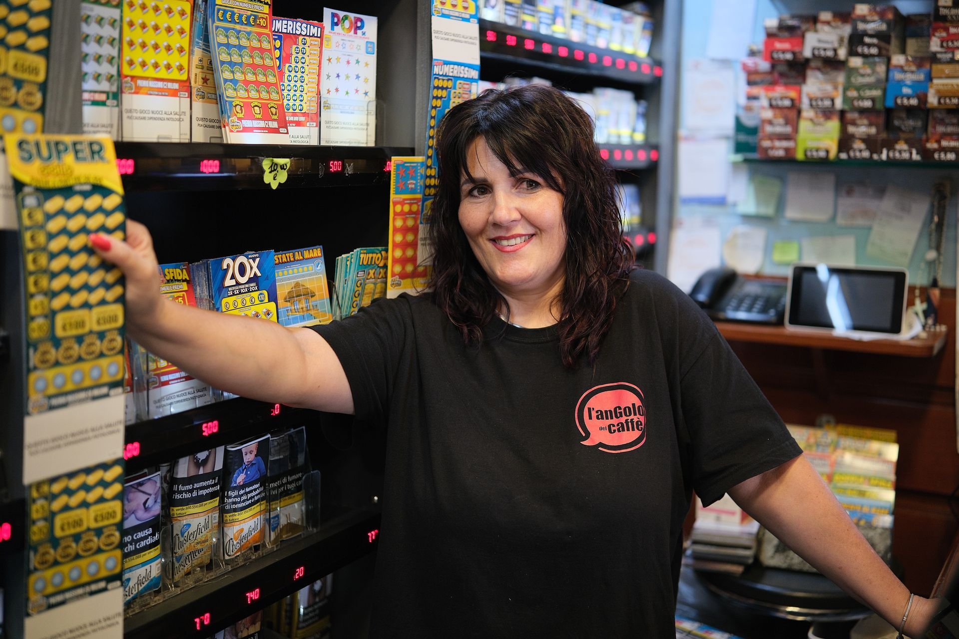 Una donna sorride, in piedi in un negozio pieno di biglietti della lotteria e prodotti per fumatori.