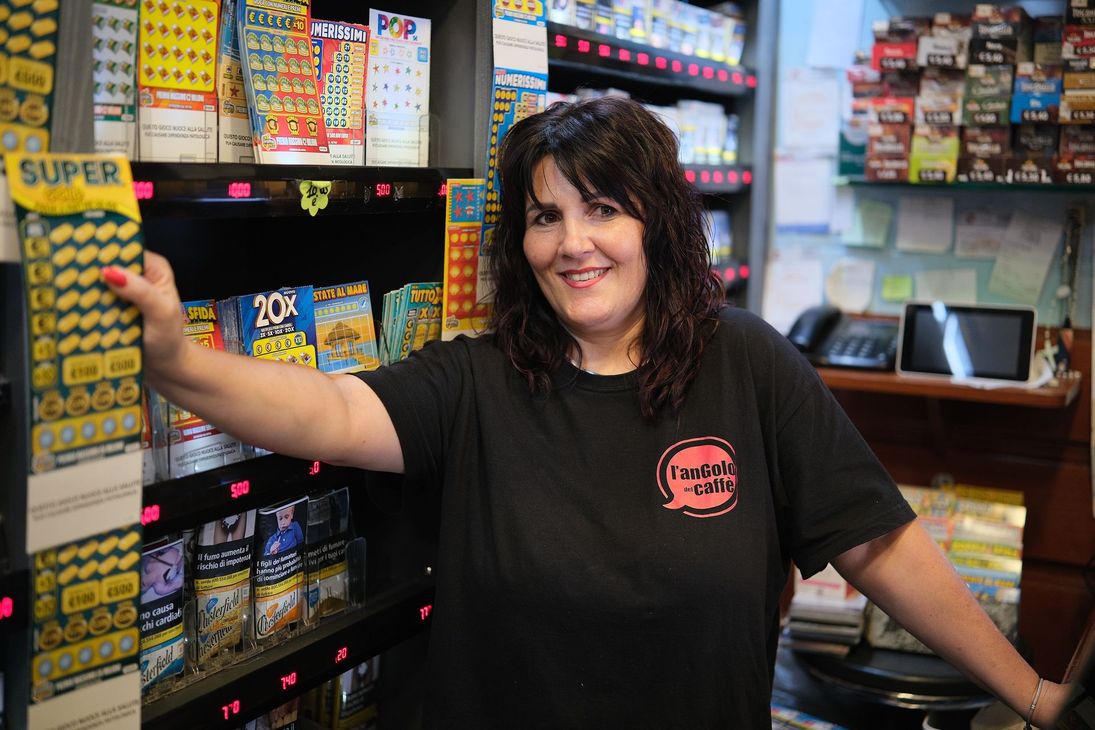 Una donna sorride, in piedi in un negozio pieno di biglietti della lotteria e prodotti per fumatori.