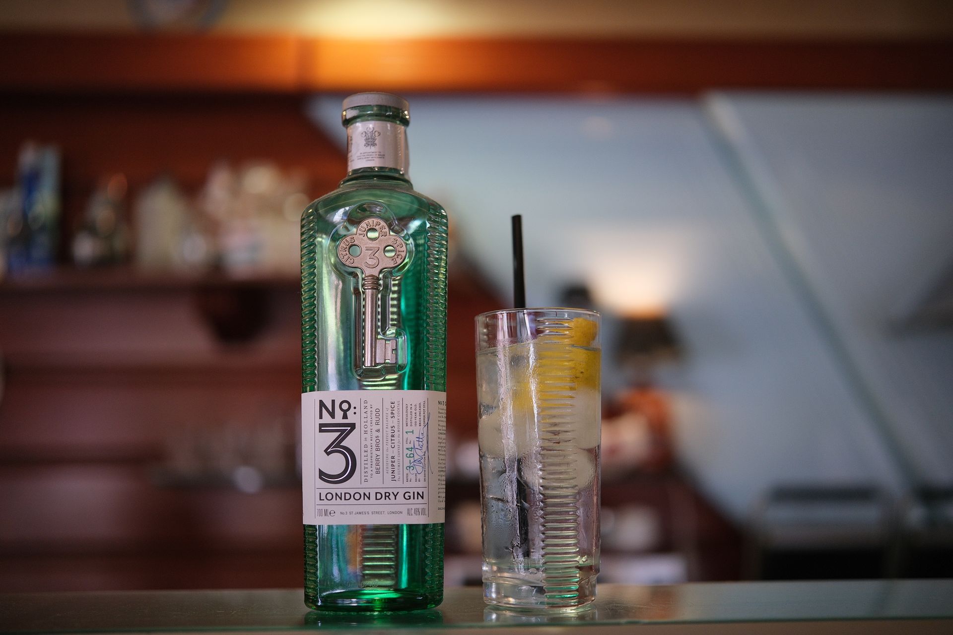 Bottiglia di London Dry Gin n. 3 accanto a un bicchiere highball con ghiaccio, limone e cannuccia su un bancone.