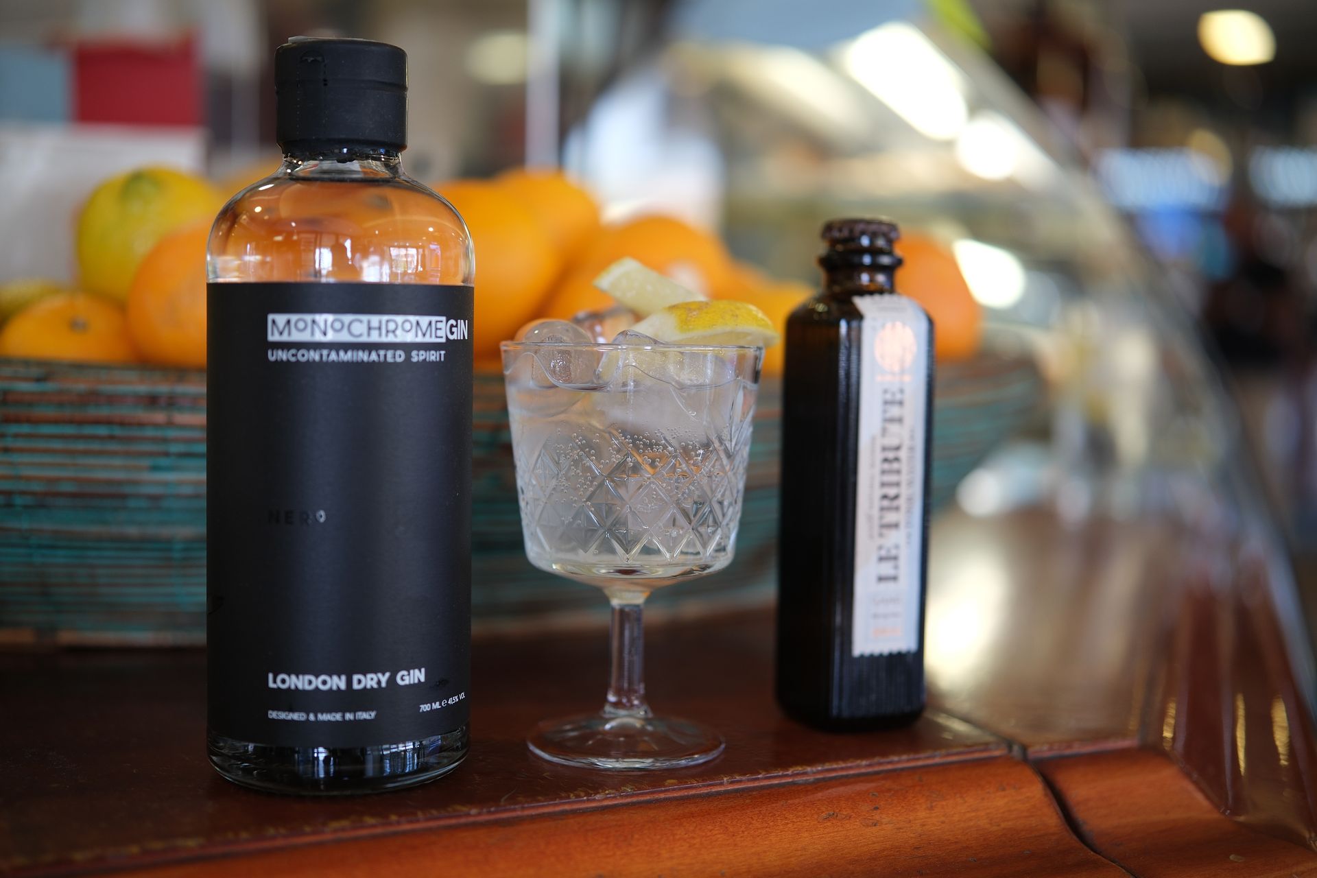 Una bottiglia di gin, un cocktail e un'altra bottiglia, posizionati su un bancone con delle arance sullo sfondo.