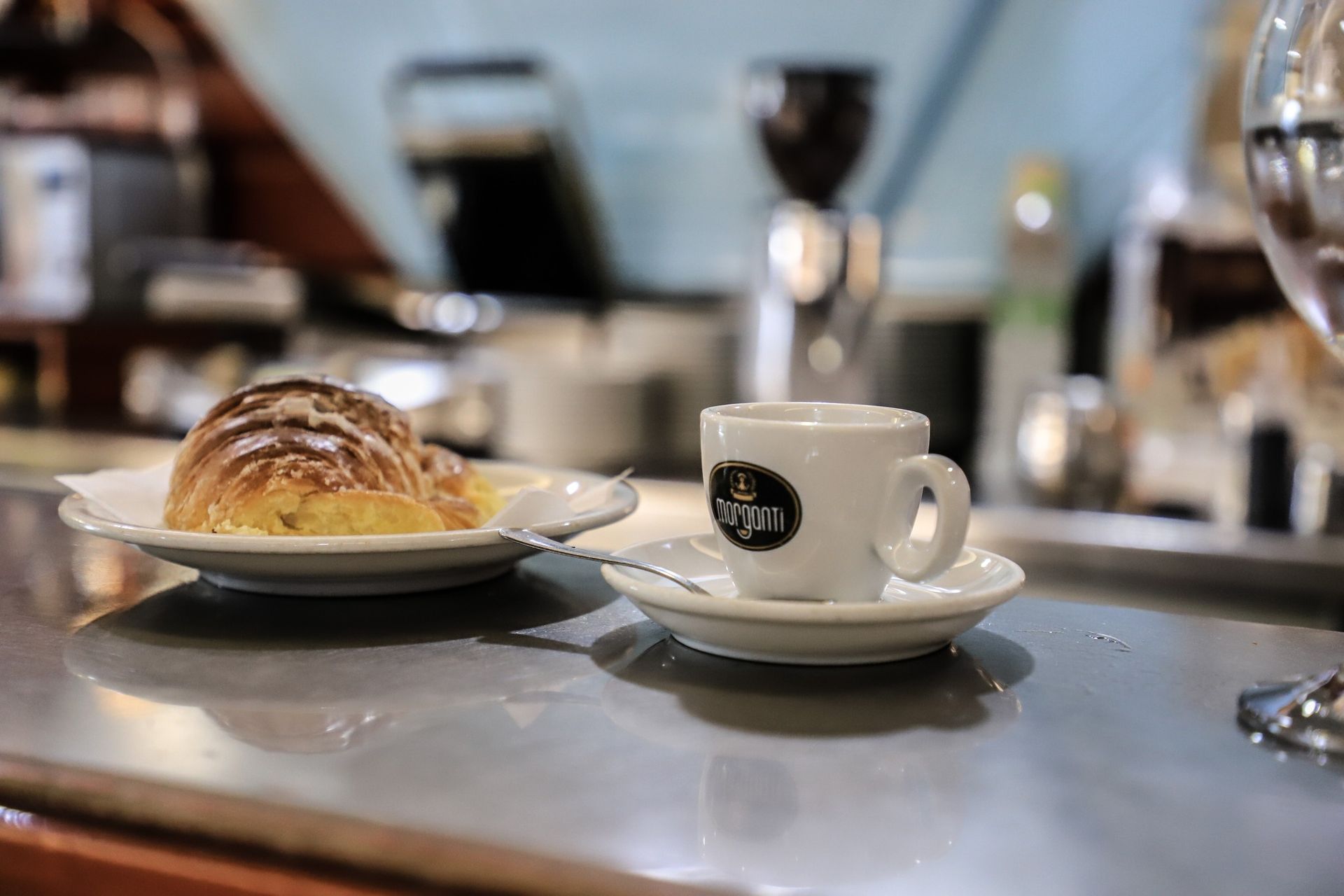 Croissant su un piatto accanto a una tazza di caffè su un bancone di metallo in un bar.