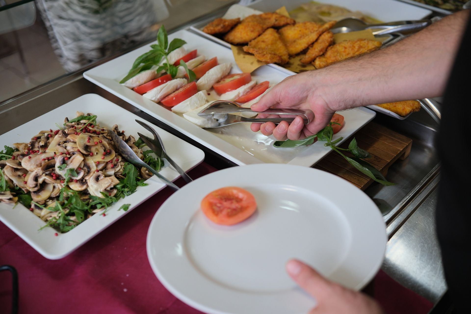 La mano di una persona usa le pinze per appoggiare una fetta di pomodoro su un piatto durante un buffet.