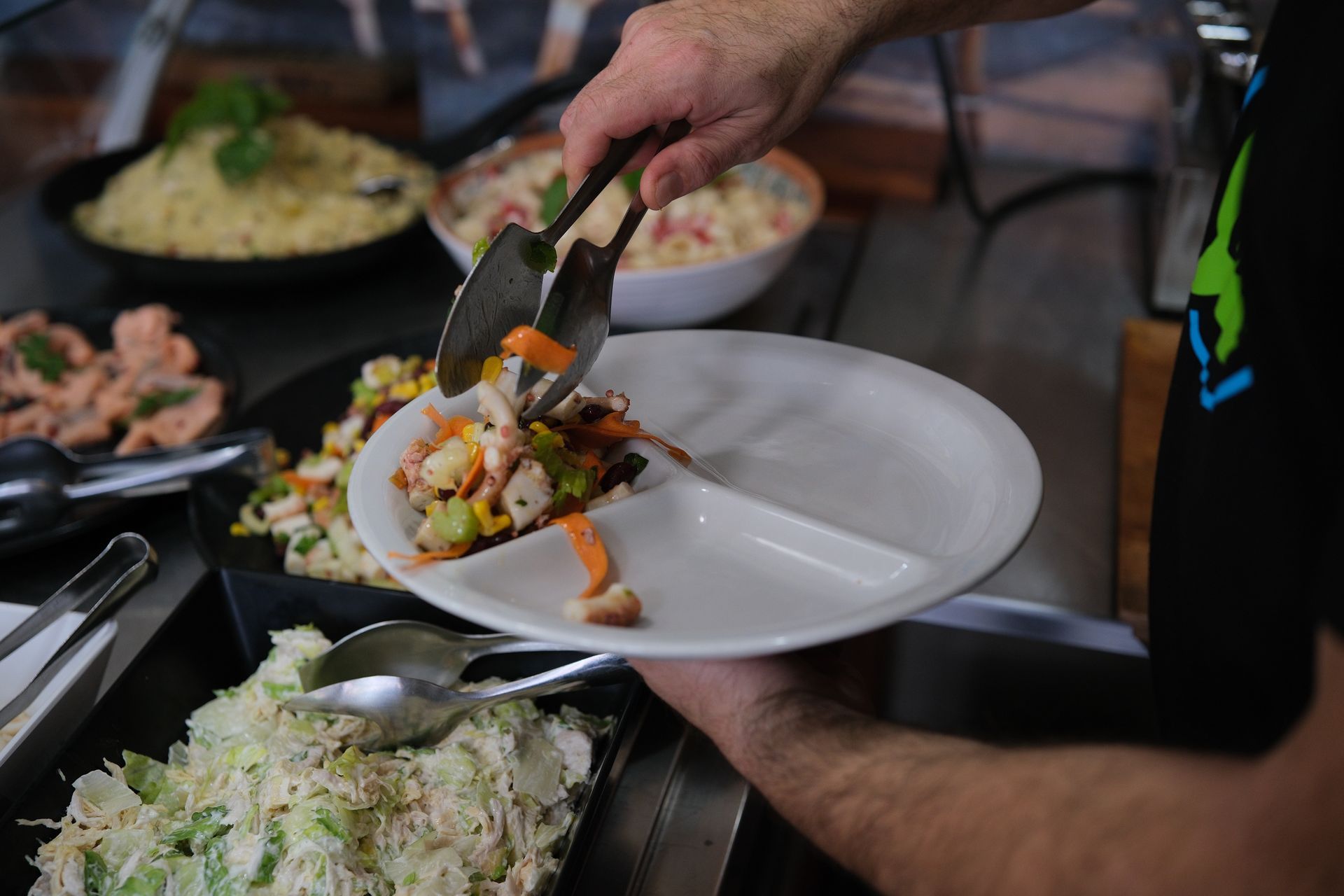 Persona che usa le pinze per disporre l'insalata su un piatto diviso in più parti durante un buffet. 