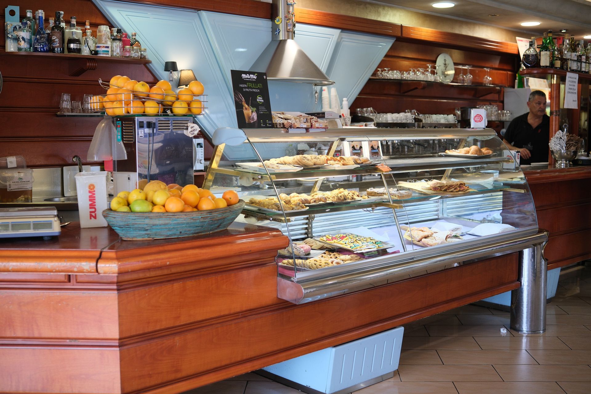 Interno di un bar con pasticcini, frutta, un barista e un bancone.