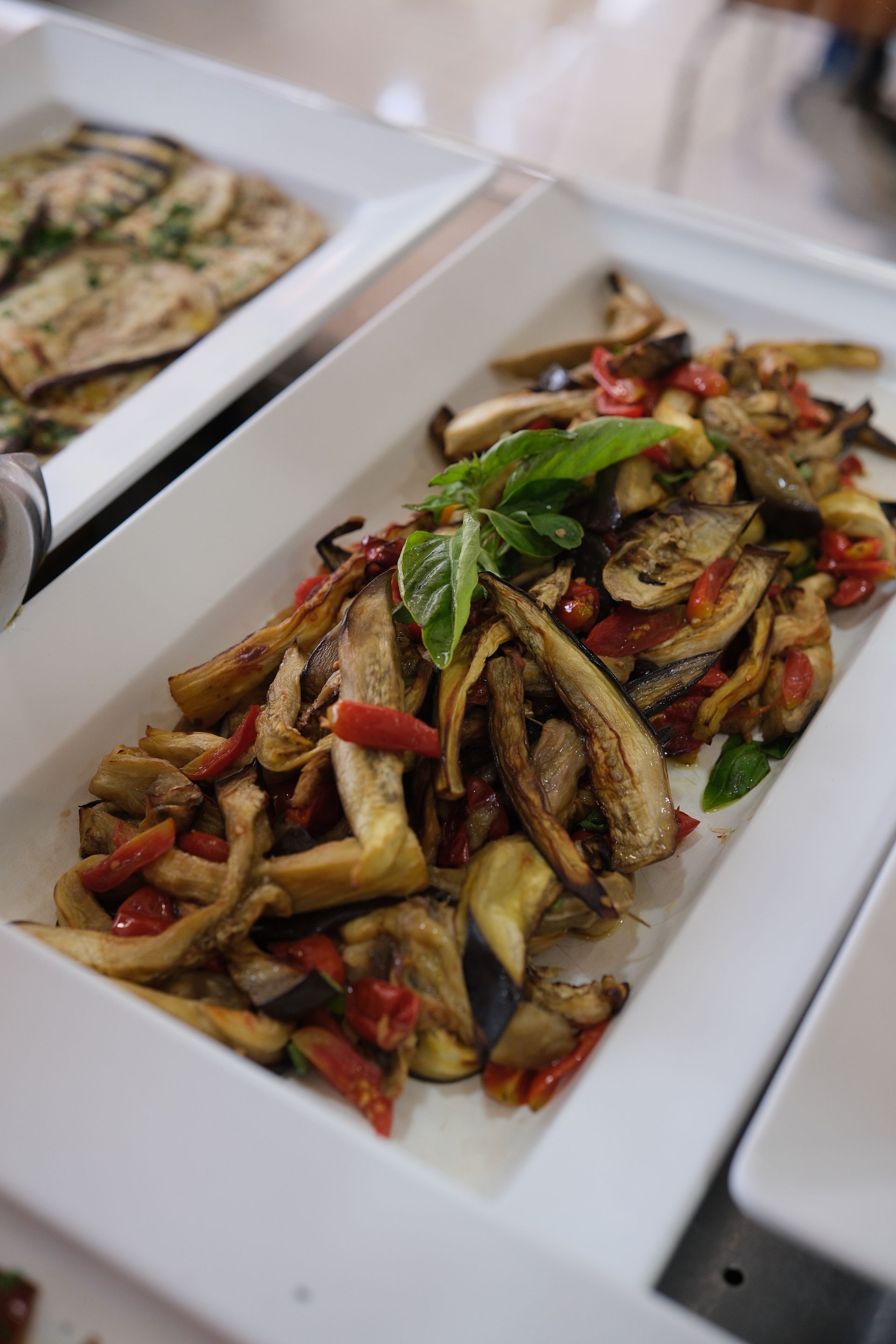Melanzane e pomodori arrostiti con basilico su un piatto bianco, parte di un buffet.