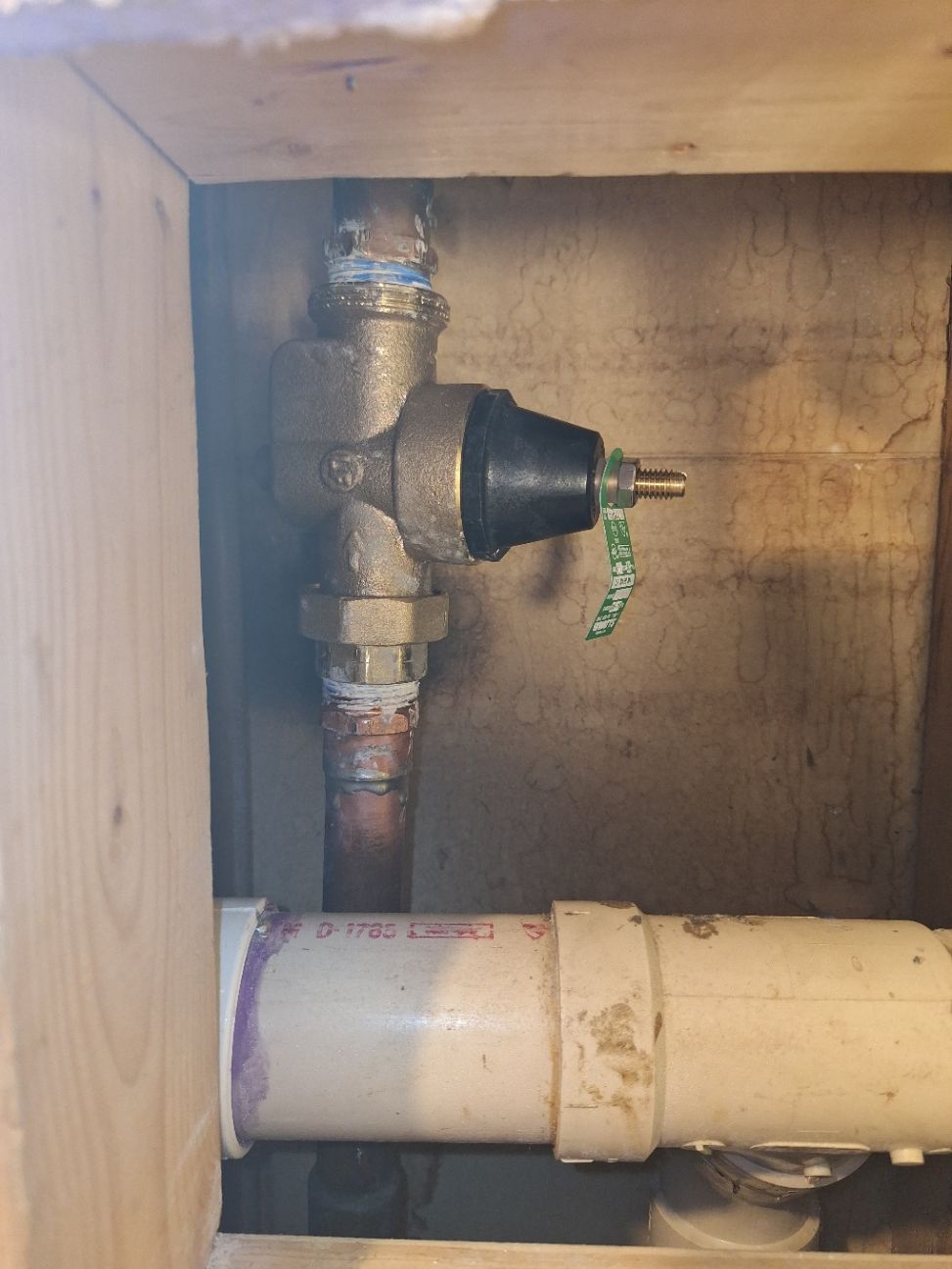 A brass water pressure regulator valve installed on copper piping within a wooden wall frame near white PVC pipes.