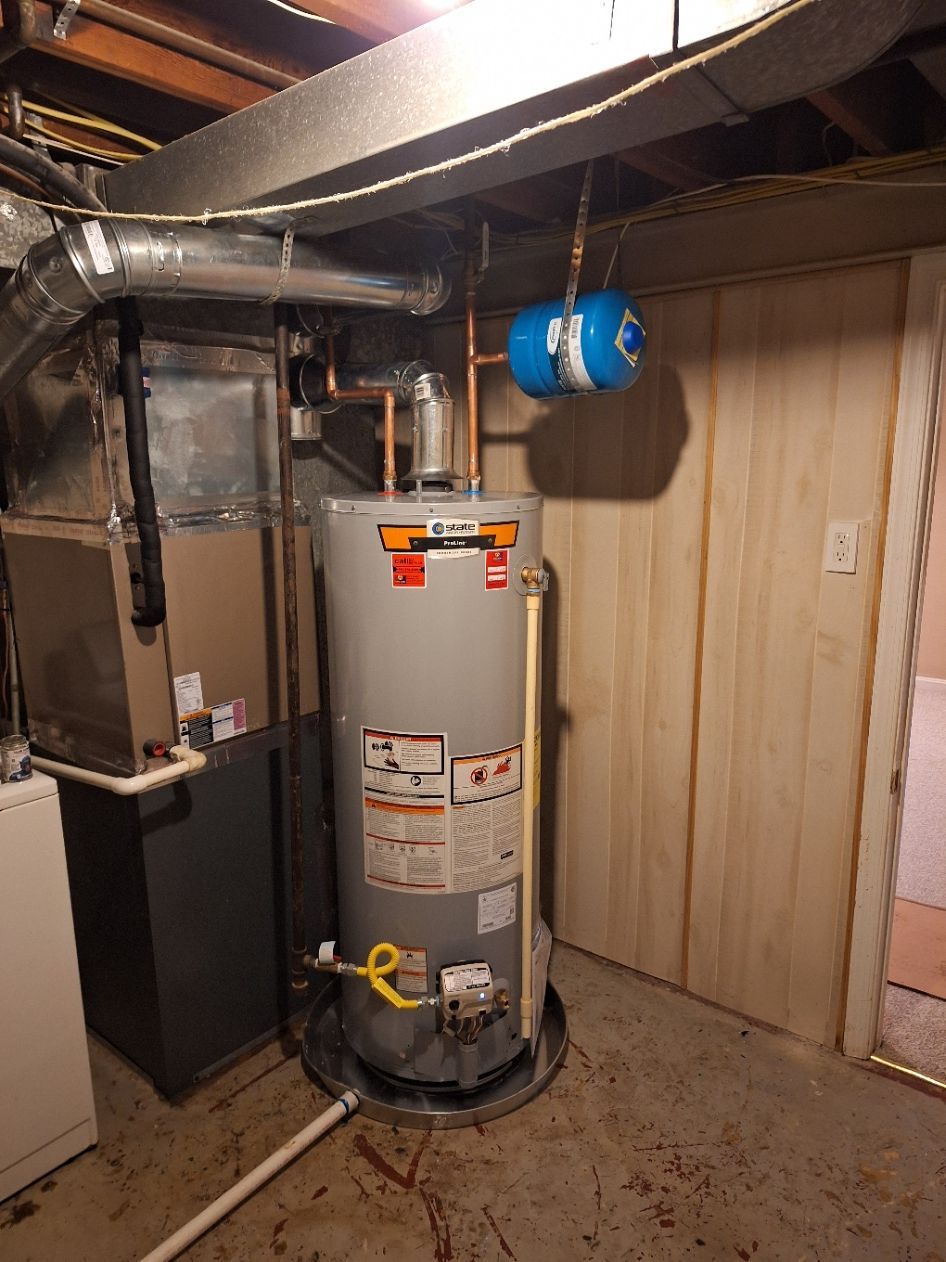 A grey water heater sits on a drain pan in a basement next to a furnace, with a blue expansion tank attached to the piping.