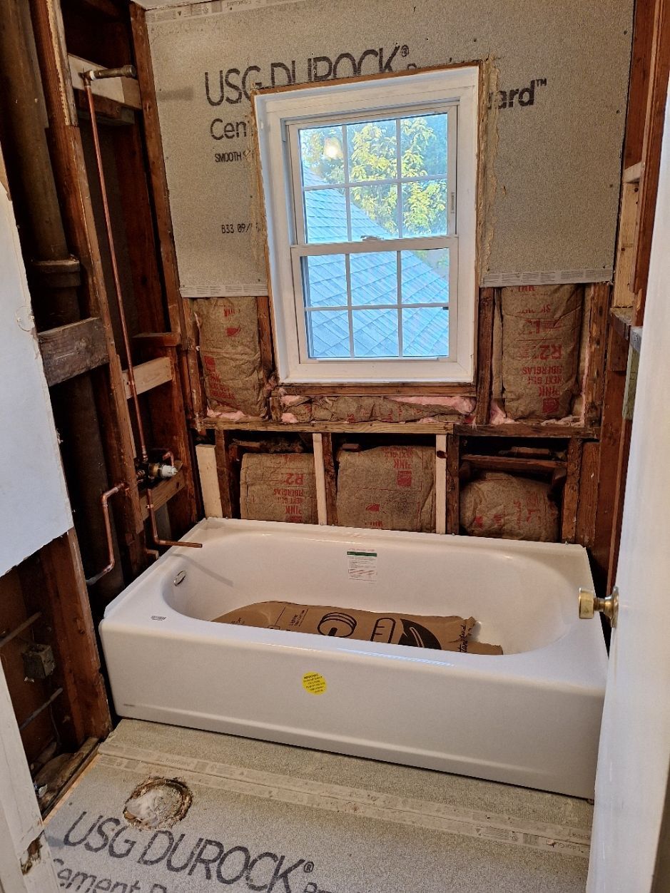 An unfinished bathroom renovation with a white bathtub installed against a wall with exposed studs, insulation, and board.