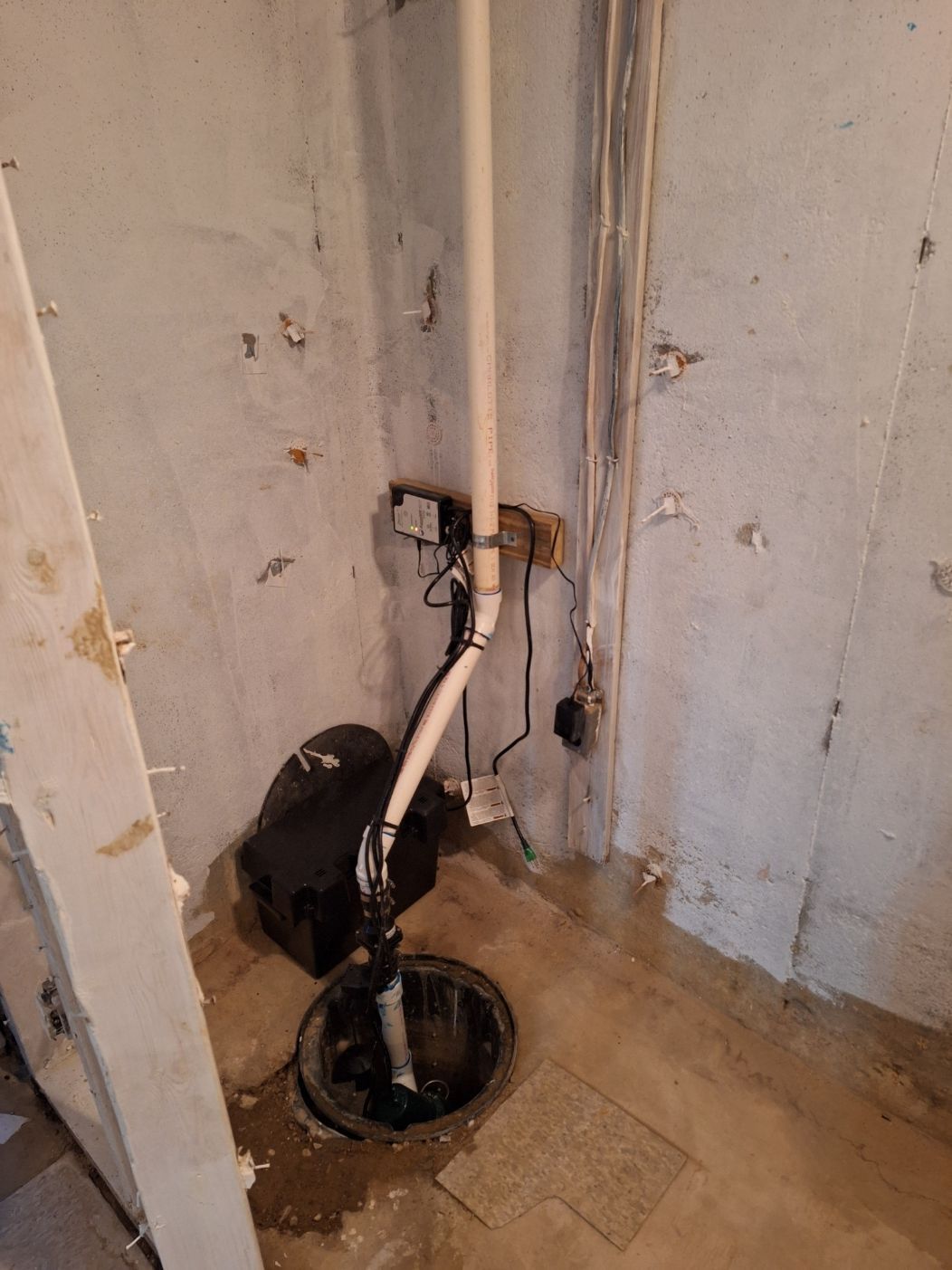 A sump pump installed in a concrete basement floor corner with a vertical white PVC discharge pipe and attached battery.