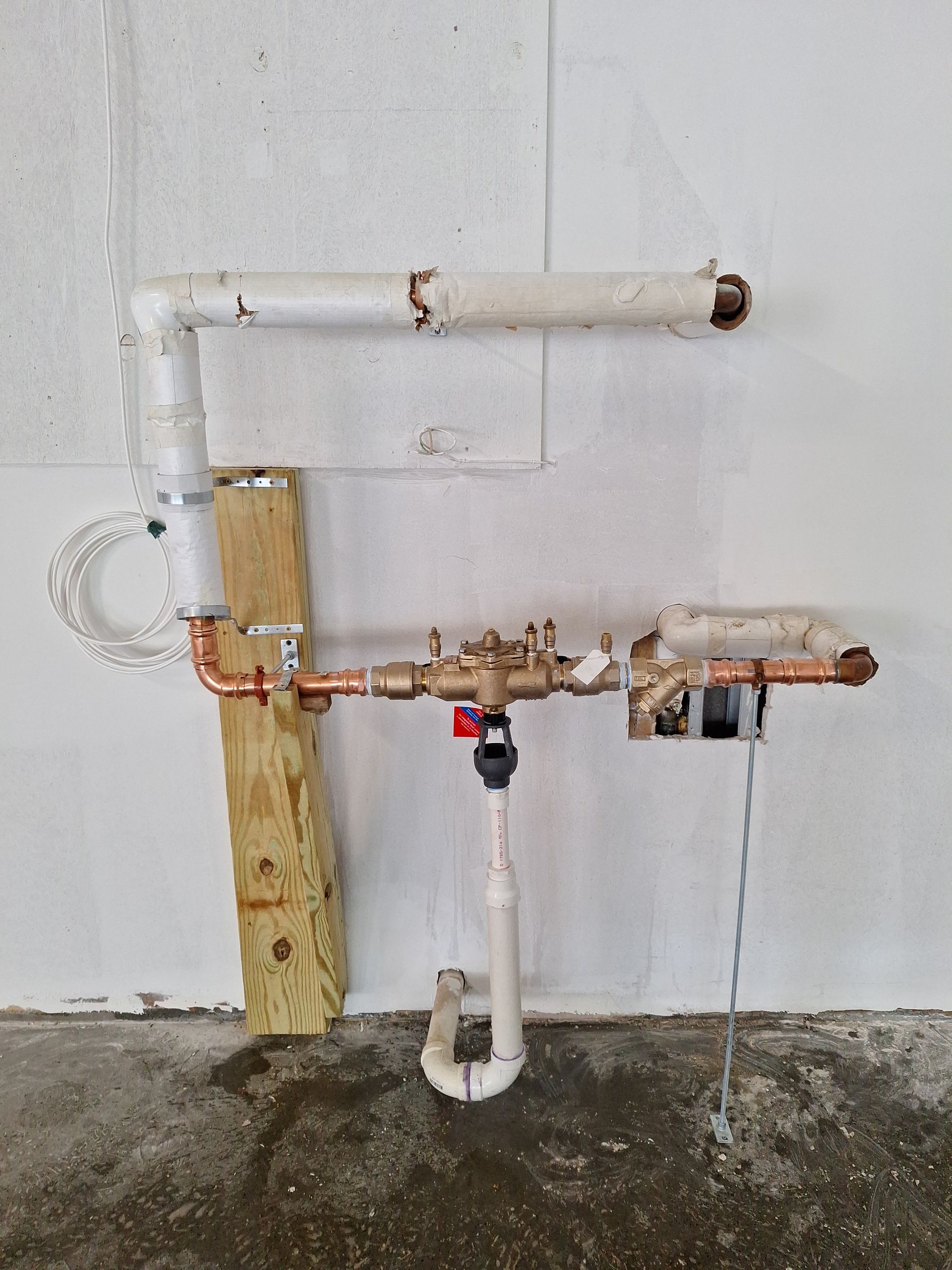 Copper and white PVC plumbing pipes installed on a white wall above a concrete floor, featuring a central brass valve.