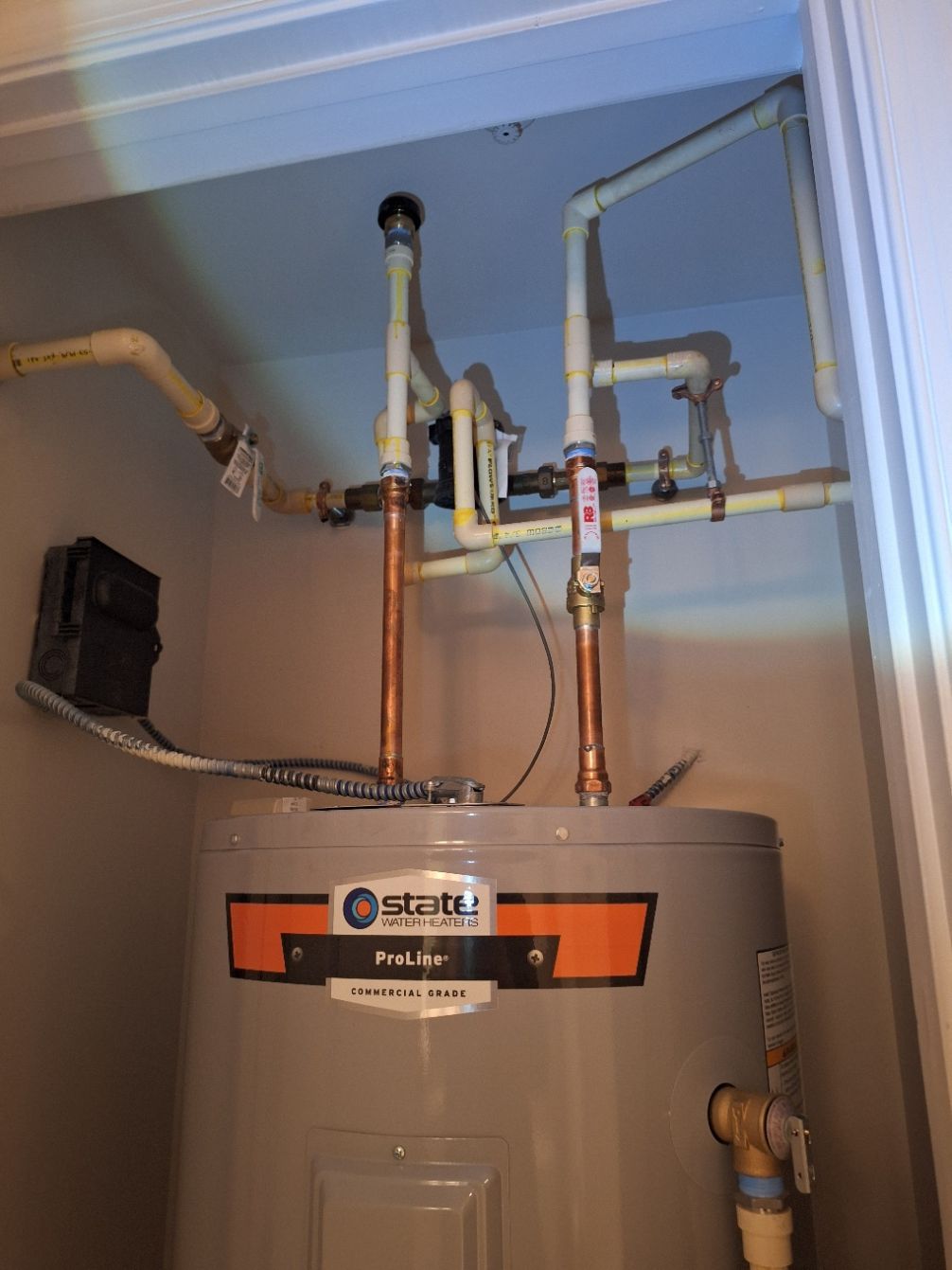 A State brand water heater with copper and beige piping in a closet setting.