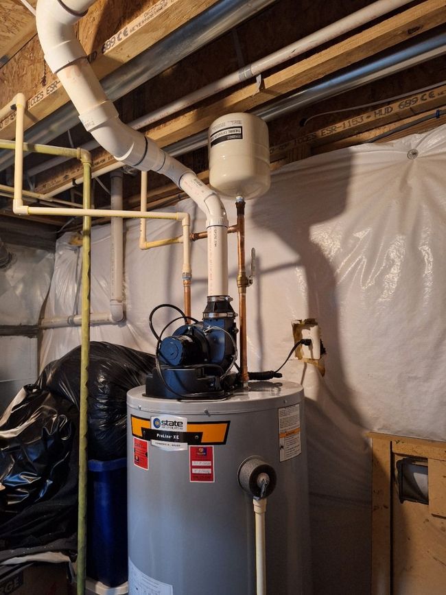 A grey water heater in a basement with white PVC exhaust piping, a small expansion tank, and a blue power vent motor.
