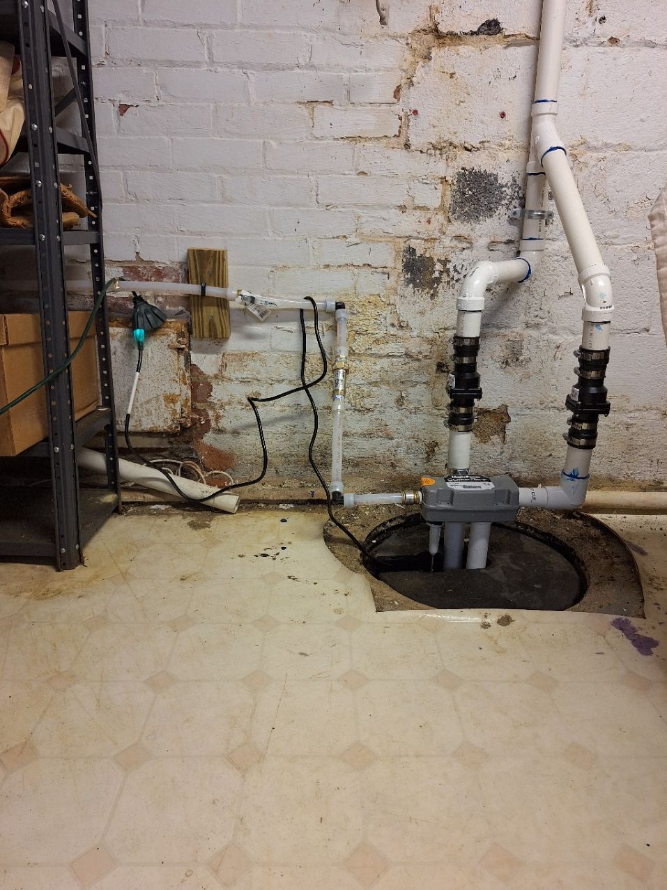 A sump pump system with two discharge pipes set into a basement floor against a brick wall.