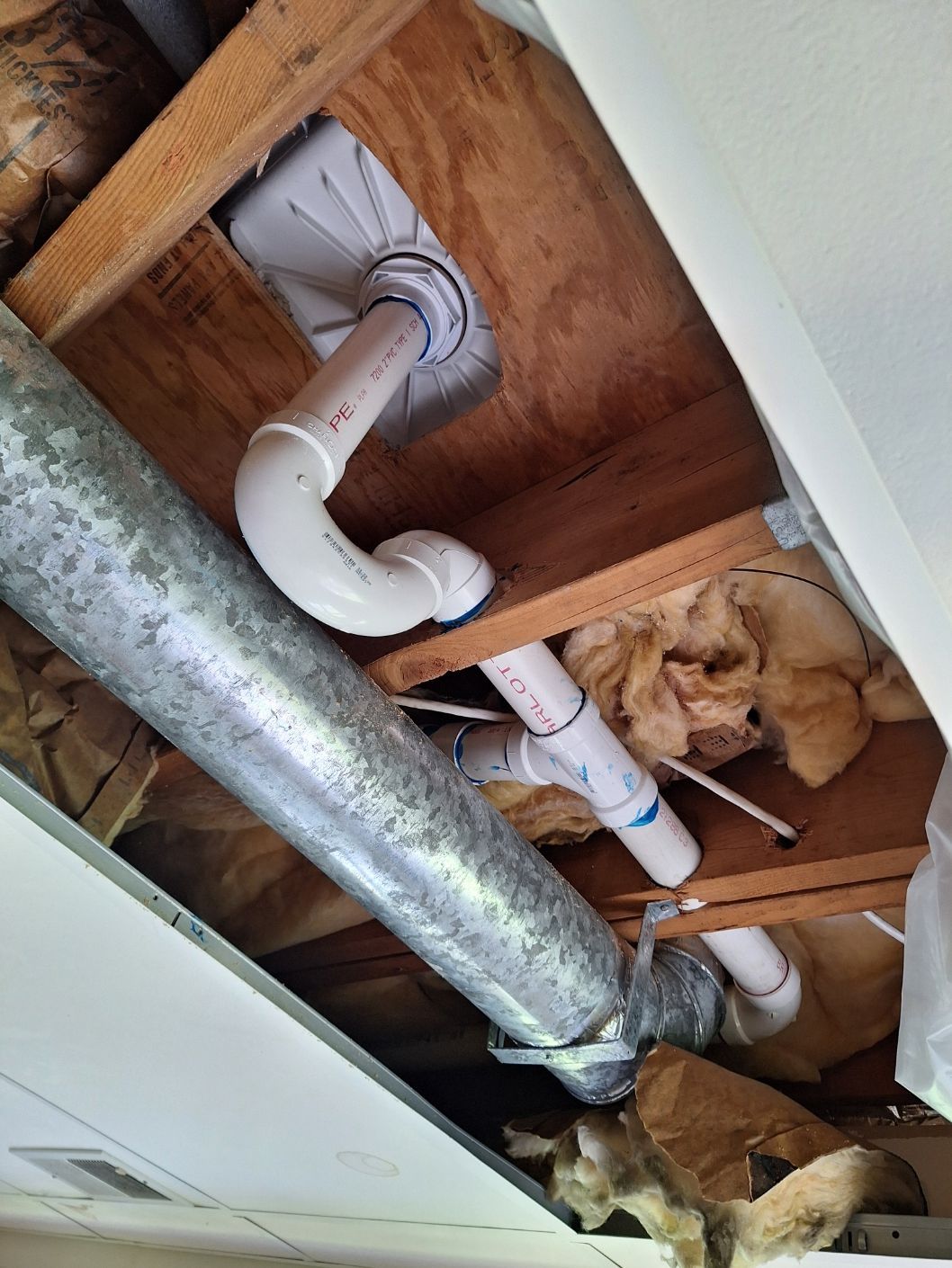 Looking up into an attic space showing white PVC plumbing pipes connected to a ceiling drain and a metal HVAC duct.