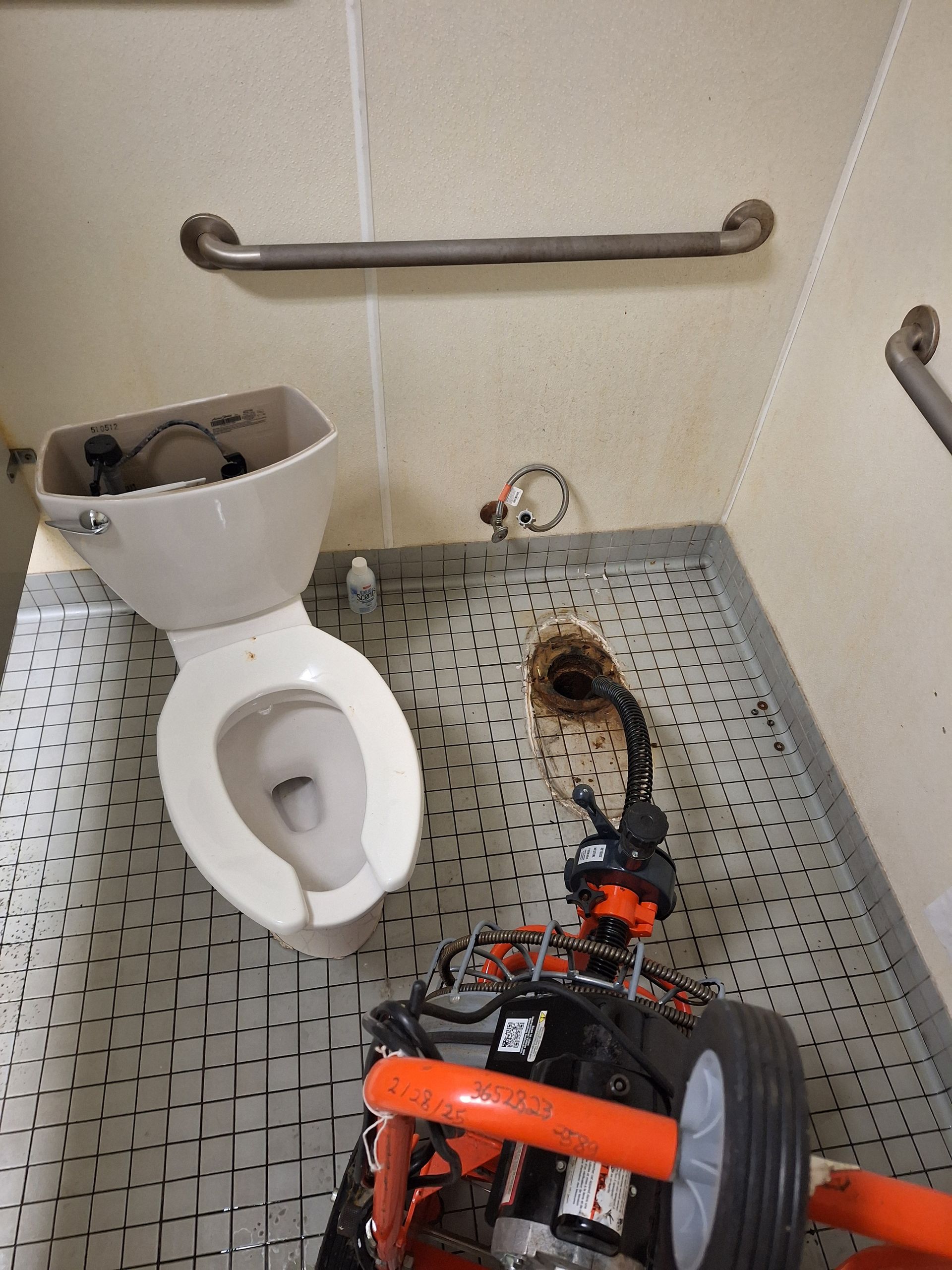 A plumber wearing grey work gloves uses an orange pipe wrench to tighten a metal drain pipe under a sink.