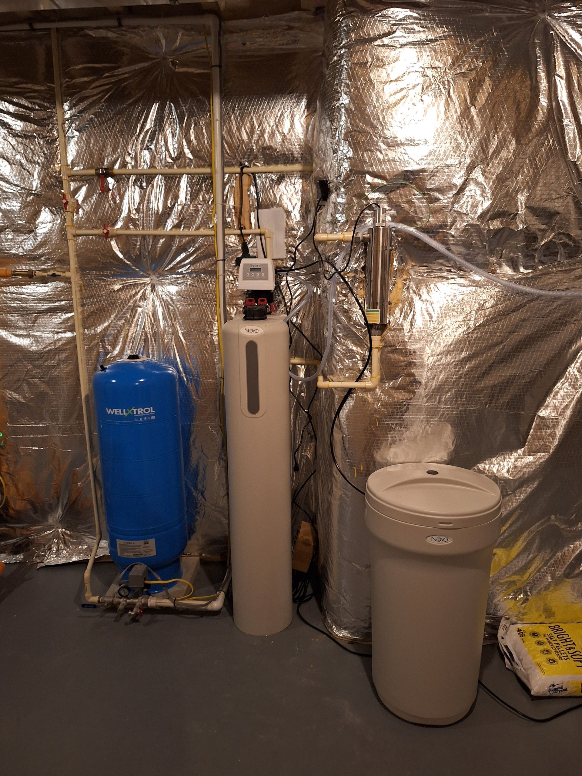 A water softener system with a blue pressure tank, beige resin tank, and brine barrel against a foil-insulated wall.
