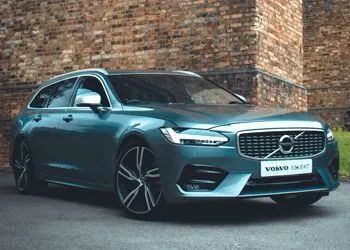 Teal Volvo V90 wagon parked in front of a brick building. Black and silver wheels.