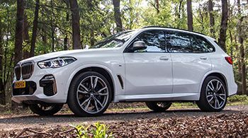 A white bmw x5 is parked on the side of a road in the woods.