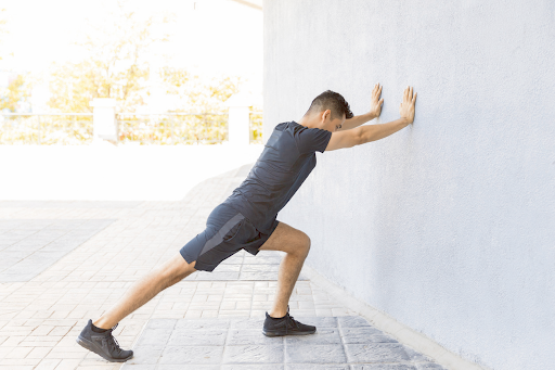 Man stretching against a white wall. He leans forward, one leg extended, hands on the wall.