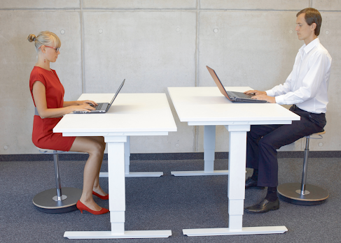 Two people seated at white adjustable desks, each with a laptop. They are indoors, and the woman is wearing a red dress.