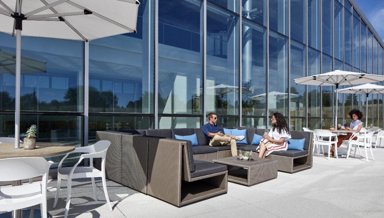 Outdoor patio with people seated on a sectional sofa, white chairs, and umbrellas; glass building in the background.