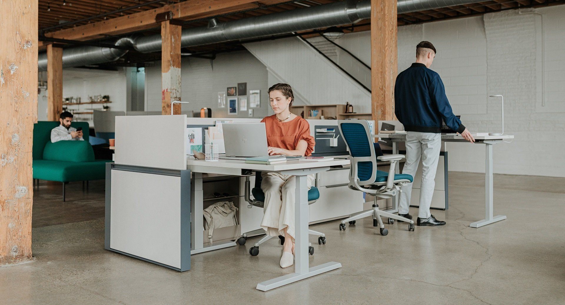 People working in a modern office. A woman uses a laptop at her desk, while others work or relax nearby.