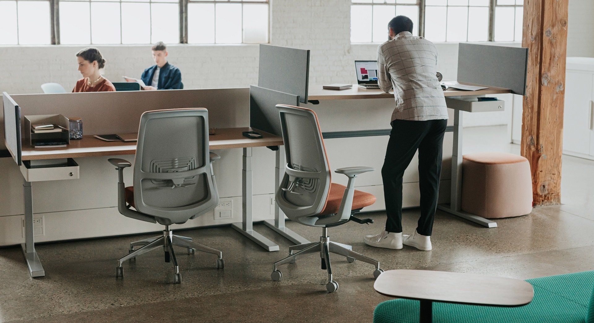 Man standing at a desk in a modern office, with two other people working nearby.
