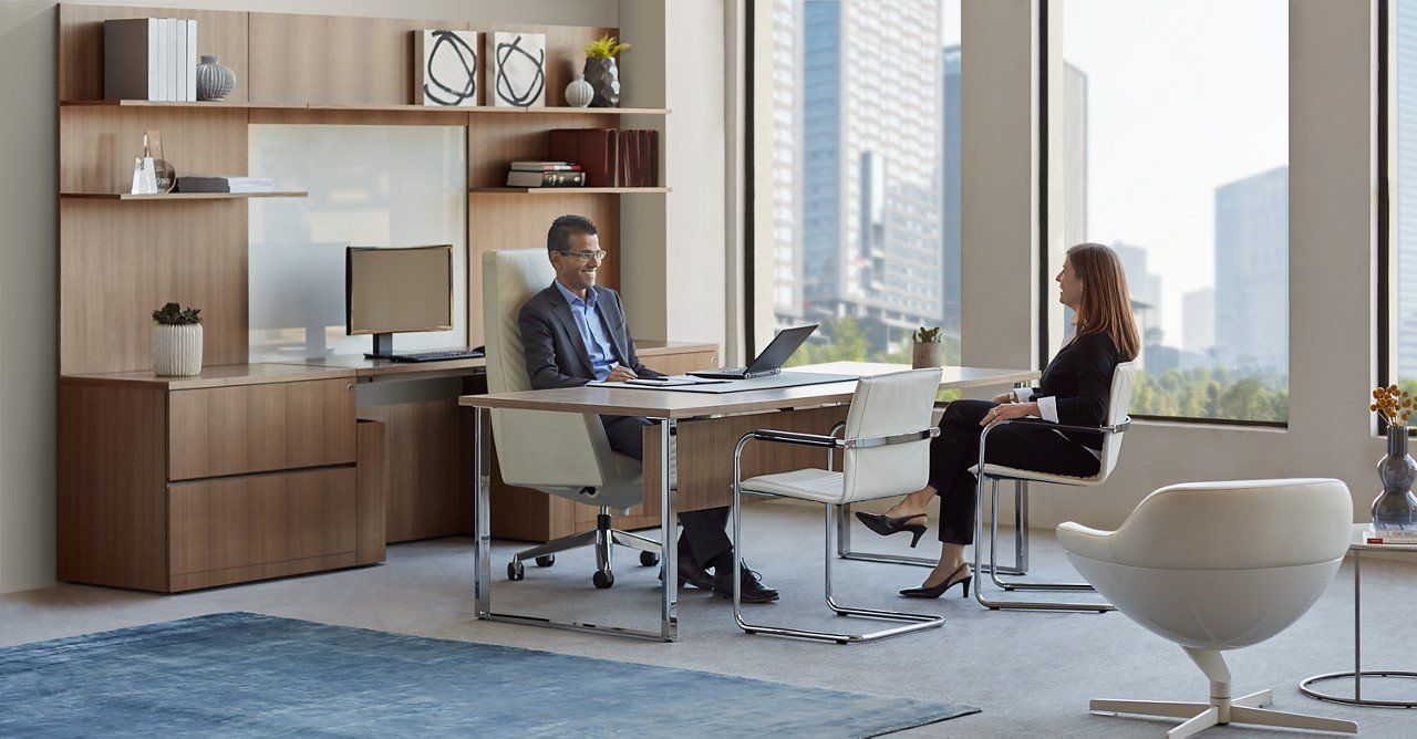 Man and woman at desk in office with city view, discussing a laptop.