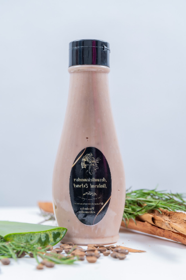 Una botella de loción de color marrón claro con una etiqueta negra reposa sobre una superficie blanca, con aloe vera, romero y granos de café cerca.