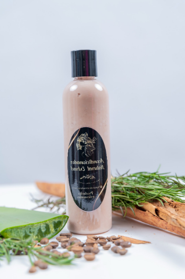 Una botella de color marrón claro de un producto para el cuidado del cabello reposa sobre una superficie blanca con aloe vera, ramitas de romero y semillas esparcidas.